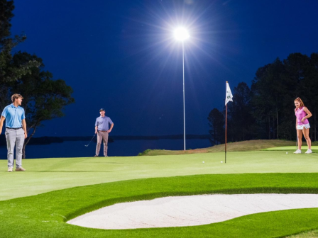 A group of three people playing golf on a well-lit green at night, with a bright moon illuminating the scene and a body of water in the background.