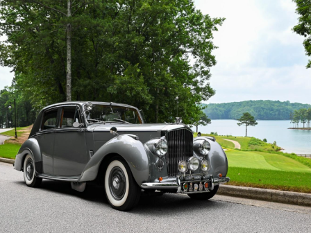 A vintage gray car is parked by a scenic lakeside with green trees and a cloudy sky in the background.