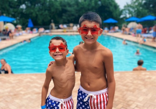 Two boys in star-spangled swim shorts and red star sunglasses smiling by a pool on a cloudy day.