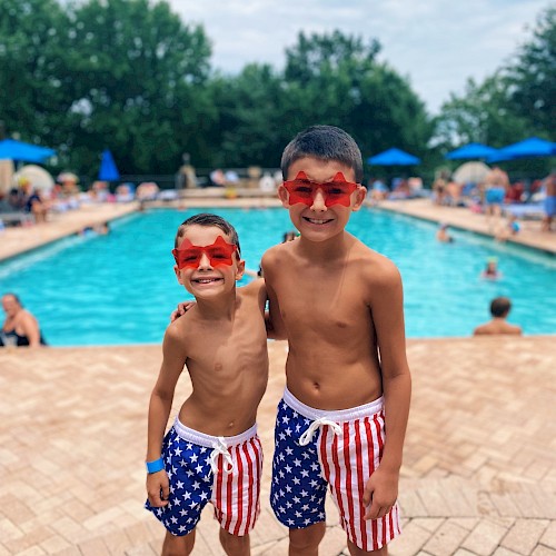 Two boys in star-spangled swim shorts and red star sunglasses smiling by a pool on a cloudy day.