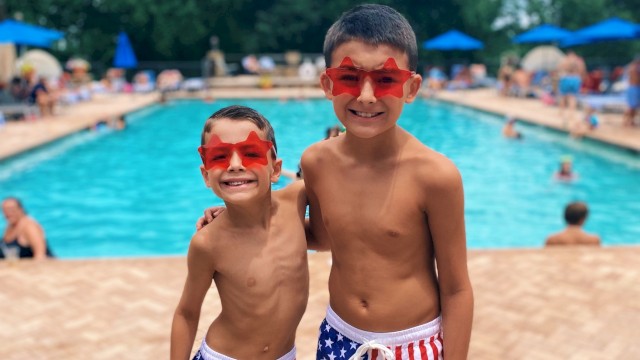Two boys in star-spangled swim shorts and red star sunglasses smiling by a pool on a cloudy day.