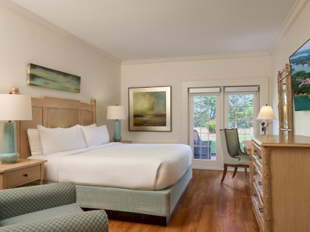 A cozy hotel bedroom with a large bed, artwork on the walls, lamps, a TV, a chair, and a balcony view of trees and water.