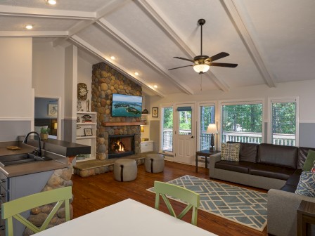 A cozy living room with a stone fireplace, large windows, a ceiling fan, and a mix of modern furniture and decor.
