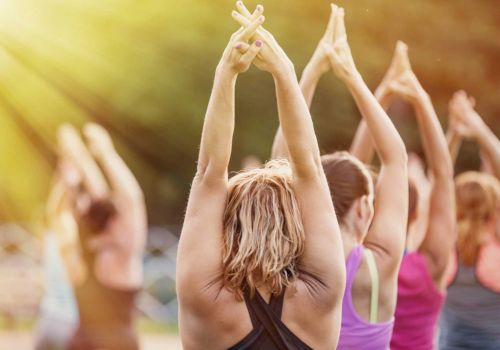 A group of people practicing yoga outdoors, raising their arms with fingers interlocked under sunlight, surrounded by nature and greenery.