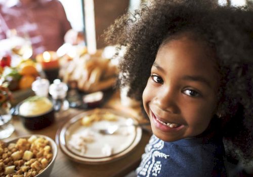A smiling child sitting at a table, surrounded by food and a festive meal setting. Other people can be seen in the background, slightly out of focus.