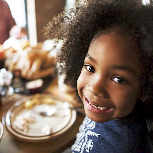 A smiling child sitting at a table, surrounded by food and a festive meal setting. Other people can be seen in the background, slightly out of focus.