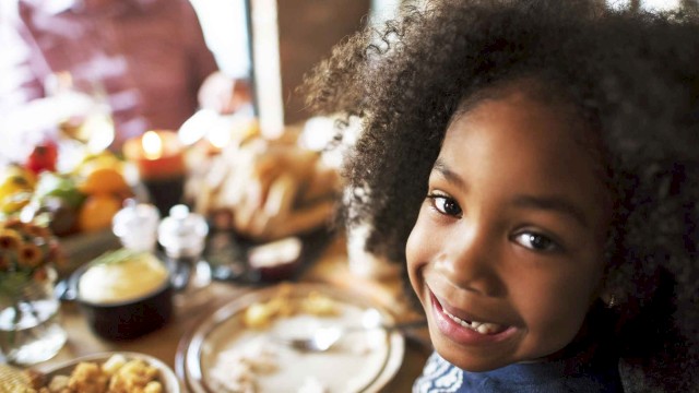 A smiling child sitting at a table, surrounded by food and a festive meal setting. Other people can be seen in the background, slightly out of focus.