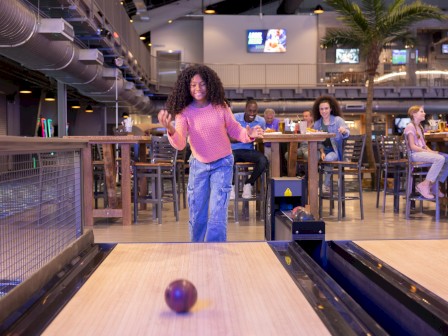 A girl in a pink sweater and jeans is bowling at an indoor alley, smiling and enjoying the game as others watch and have fun.