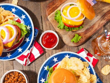 A patriotic American meal with cheeseburgers, potato chips, watermelon, baked beans, and soda on a rustic wooden table.