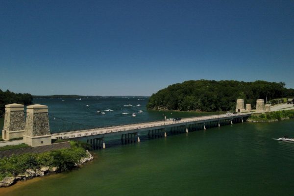 A scenic view of a bridge over a body of water, with boats in the background and lush green trees on either side of the bridge.