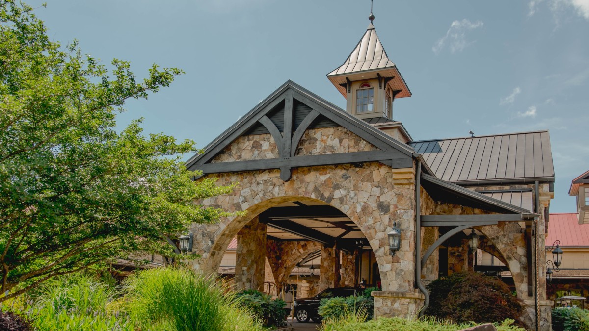 A beautiful stone building with a steep roof, turret, lush landscaping, and a car parked under the archway on a sunny day.