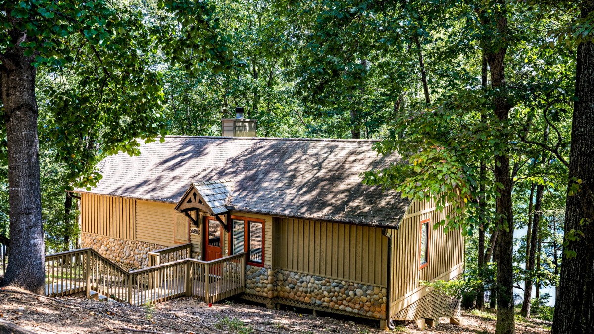 A cozy house made of wood and stone is nestled among tall trees in a dense forest by a lake or river.