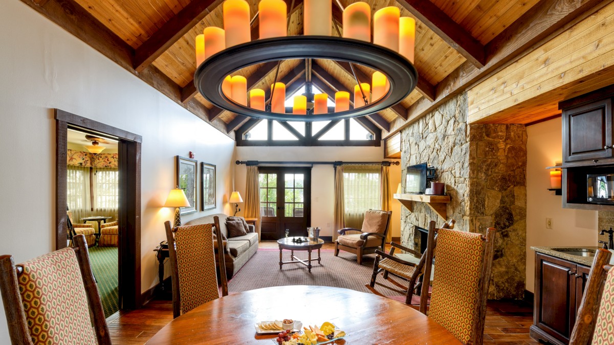 A cozy living and dining area with a wooden ceiling, stone fireplace, large chandelier, and inviting furniture in a warm, rustic setting.