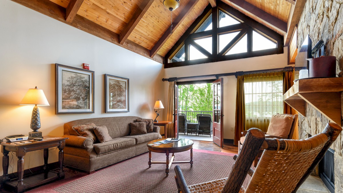 A cozy living room with a high wooden ceiling, large windows opening to a balcony, comfy chairs, a sofa, and warm lighting.