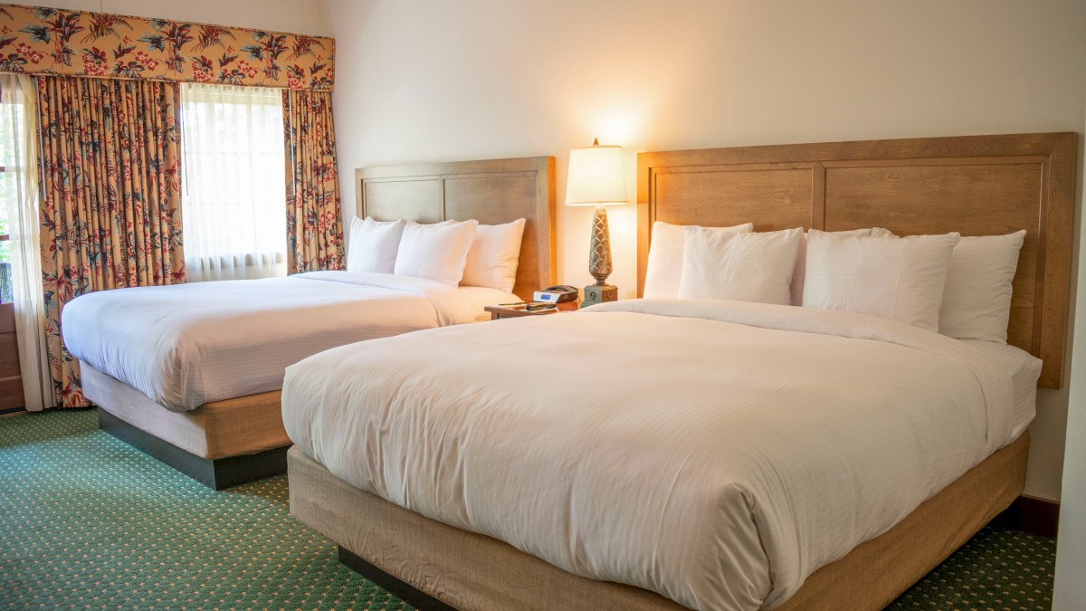 Two neatly made hotel beds with white linens and pillows are in a room with floral curtains, a lamp, and a beige carpet.