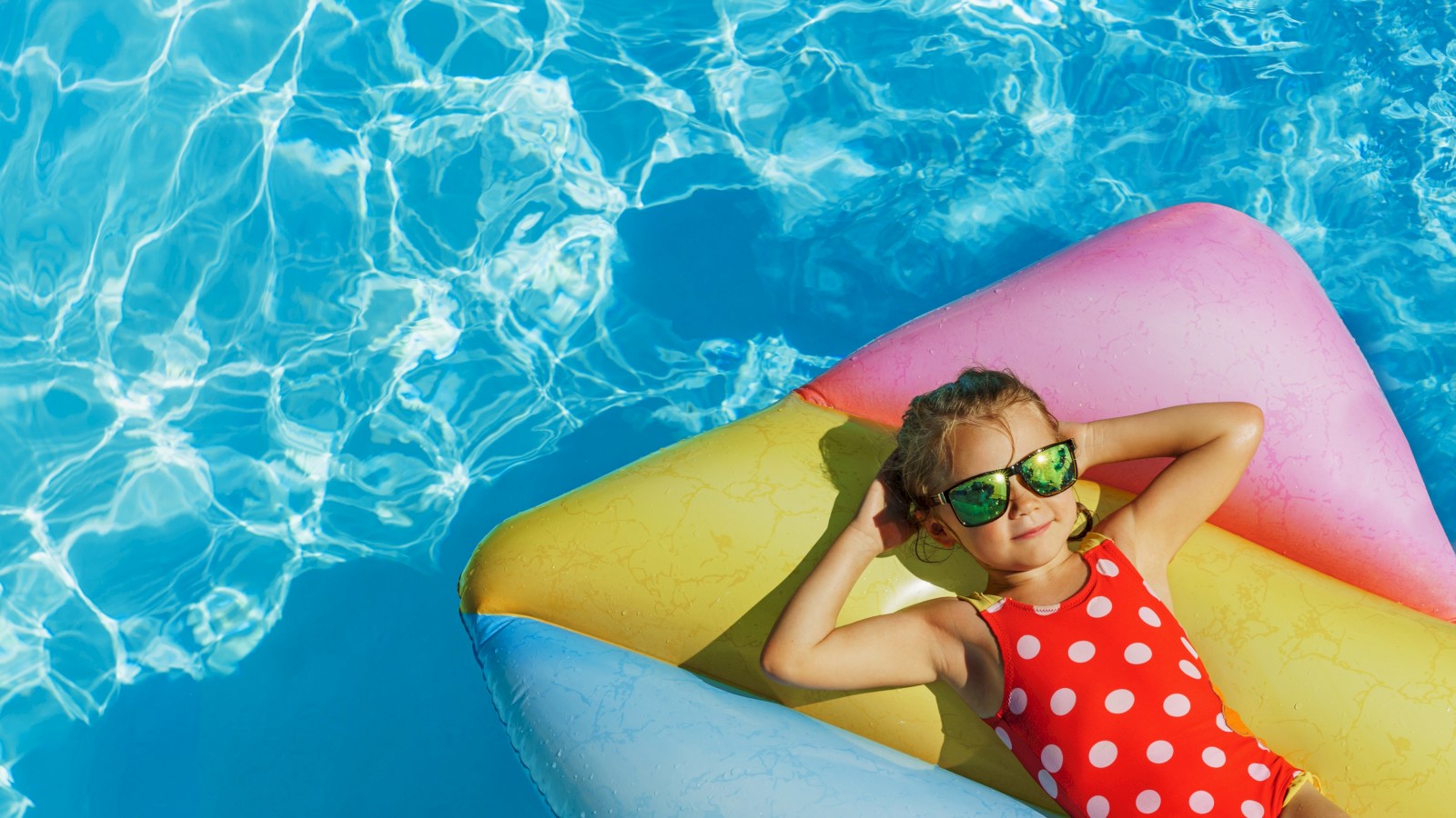 A girl in a red polka dot swimsuit with sunglasses is relaxing on a colorful float in the pool on a sunny day.