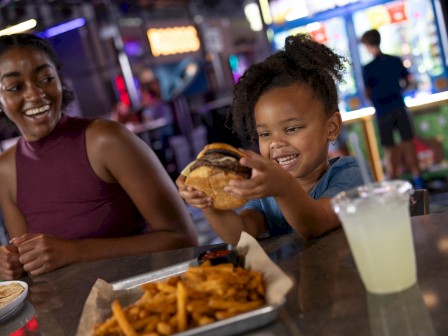 A young girl excitedly holds a burger while smiling, sitting with a woman at a lively arcade or entertainment venue.