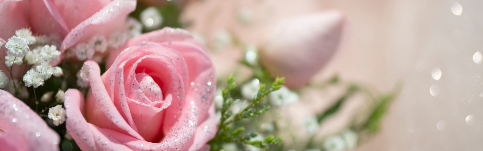 A close-up of pink roses with white baby's breath and greenery, decorated with glitter, on a soft, sparkling background.