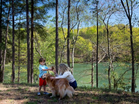 A woman, a boy, and a dog are enjoying a sunny day by a forested lake.