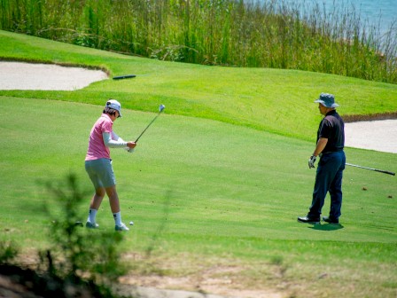 A person is preparing to hit a golf ball while another person watches on a sunny golf course with sand traps and water in the background.