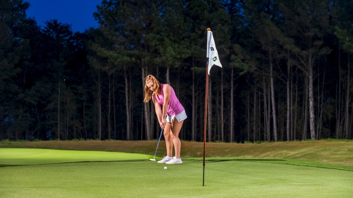 A woman in a pink top and white shorts is puttingt on a golf course at dusk near a flagstick.