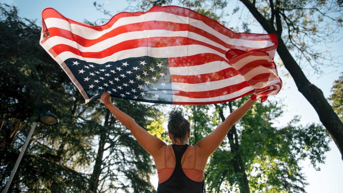 A person with curly hair holds up an American flag in a park with trees and blue sky.
