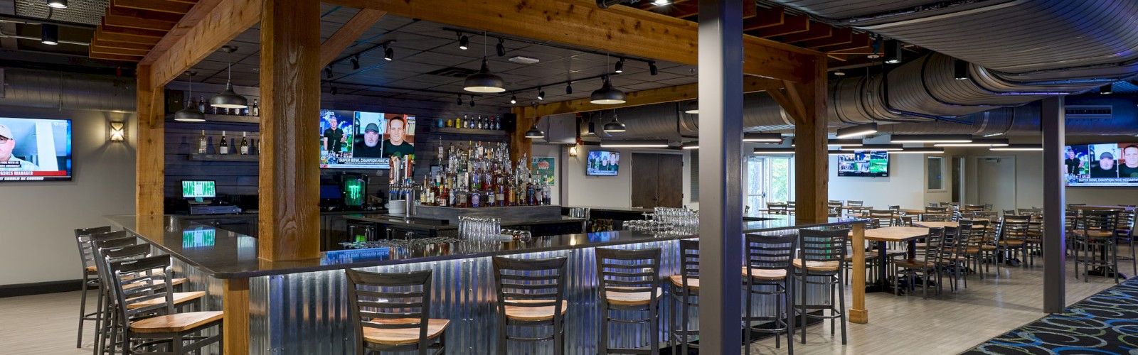 A modern sports bar with a long central counter, wooden beams, high chairs, multiple TVs, and cozy seating around the perimeter.