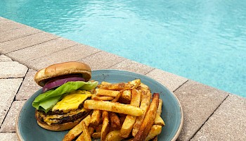 A plate with a cheeseburger and fries is placed beside a swimming pool surrounded by lounge chairs and umbrellas.