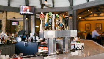 A bar scene with various beer taps, a TV in the background, and a few patrons in a spacious and welcoming setting.