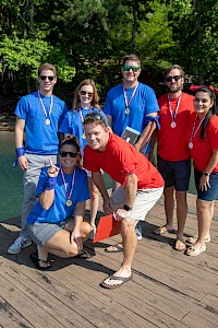 A group of seven people celebrating on a dock, with medals, outdoors by a lake and trees, on a sunny day, possibly after a race or event.