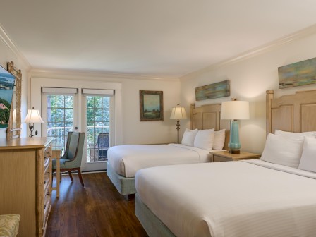 This is an image of a bright, cozy hotel room with two large beds, wooden furniture, a door leading to a balcony, and scenic views outside.