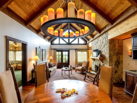 A cozy living room with wooden ceiling, stone fireplace, yellow curtains, and a chandelier with orange candles.