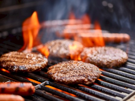 Burgers and sausages cooking on a grill with flames rising.