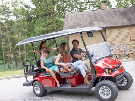 A family enjoying a ride in a red golf cart with four seats, smiling and chatting outdoors near a house with trees in the background.