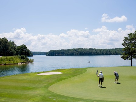 Two golfers are on a lush green golf course near a lake, with a boat visible on the water under a partly cloudy sky.