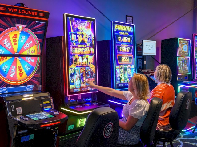 Two women are sitting in front of brightly lit arcade slot machines in a game room.