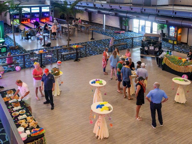 People are gathered in a decorated indoor space with food, games, and leis, suggesting a party or celebration with tropical decorations.