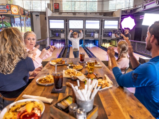 A family celebrates a bowl victory at a bowling alley, enjoying food and drinks, with a cameraman filming their fun moment.
