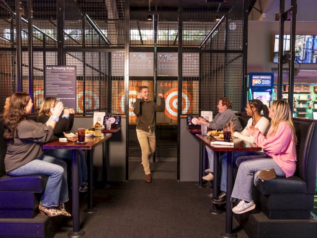 A group of people sitting at tables in a laser tag restaurant, watching a person walk in and interact, with laser tag equipment visible.