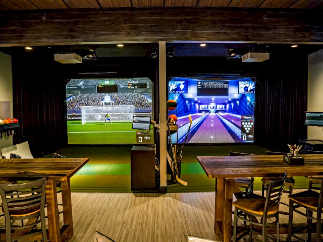 This is an indoor gaming room with two screens displaying virtual sports games: soccer and bowling.