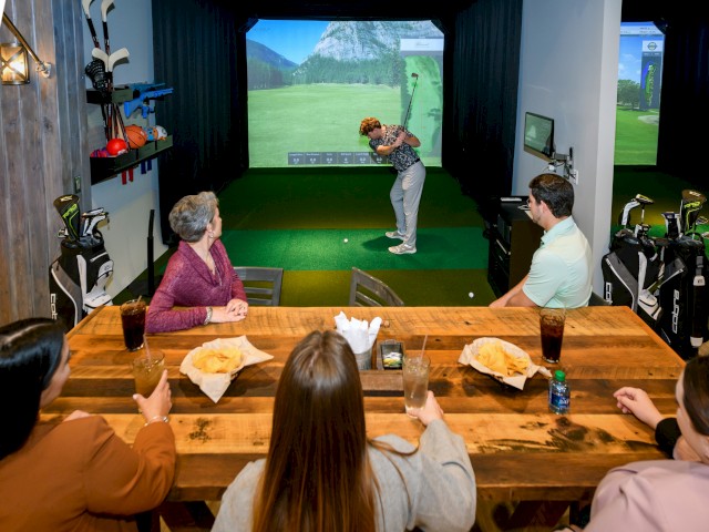 People watching a virtual golf game at a cozy indoor venue, with drinks and snacks at the table.