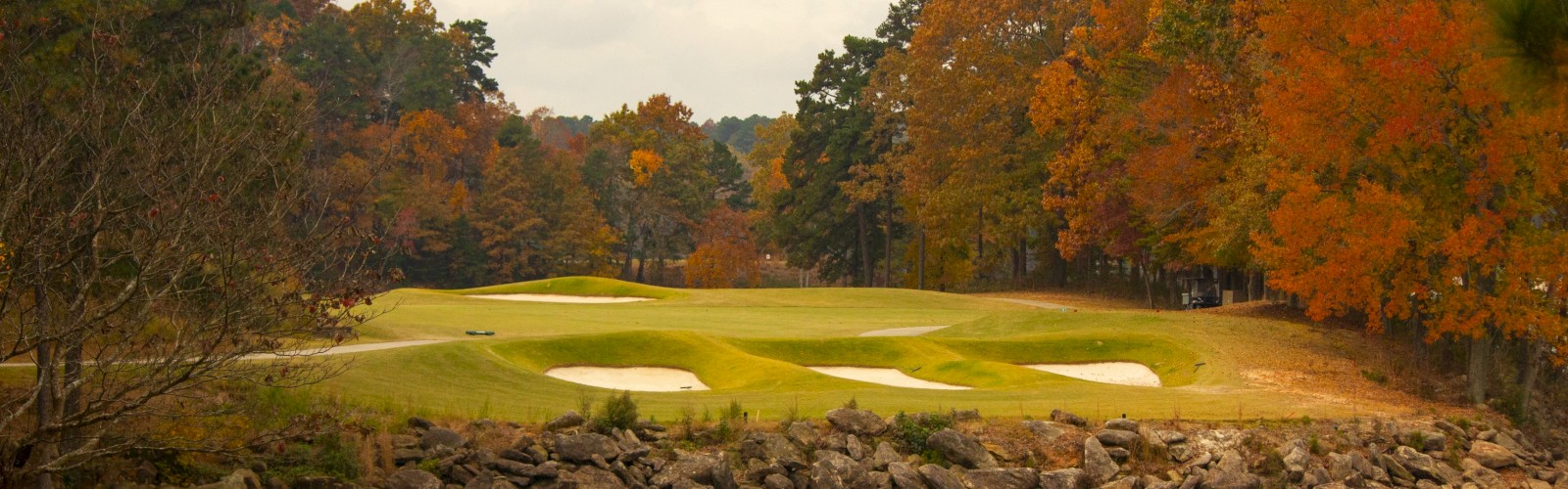 A serene autumn golf course with a lush fairway, sand traps, and vibrant foliage, bordered by a small pond under a cloudy sky.