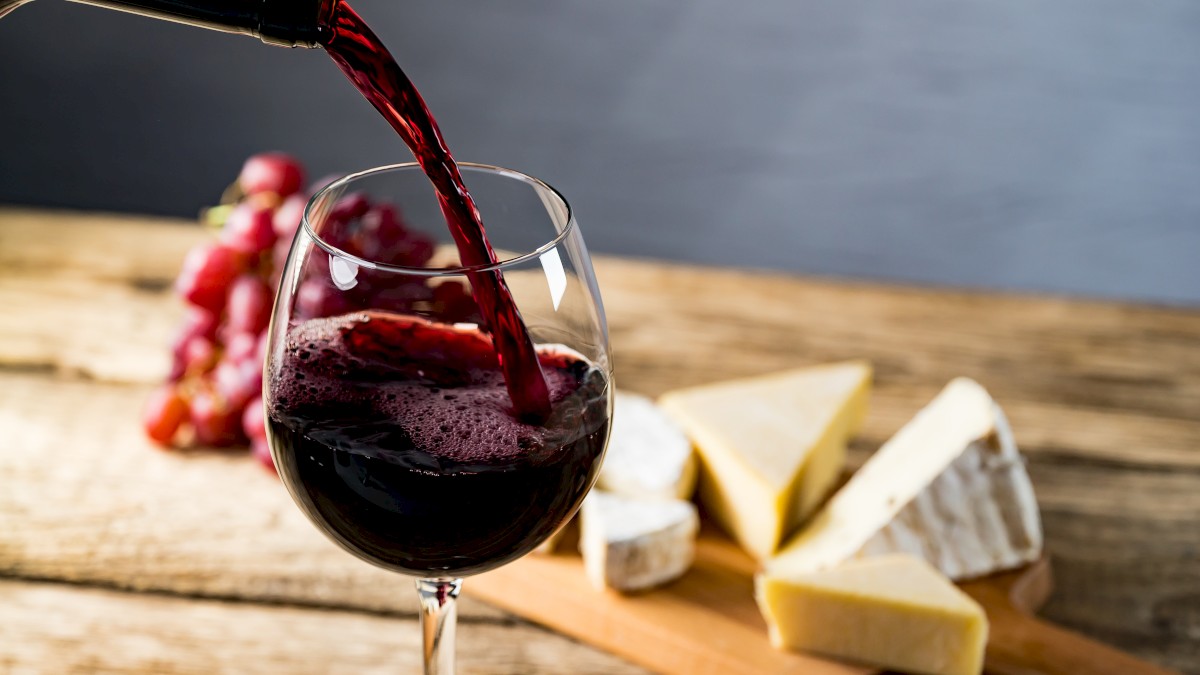 A glass of red wine being poured, with cheese wedges on a board and grapes in the background on a wooden table.