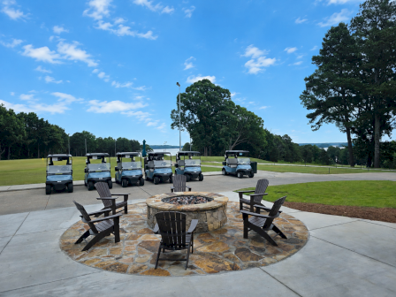 Golf carts parked on a green field with a firepit surrounded by chairs in the foreground and a blue sky with scattered clouds.