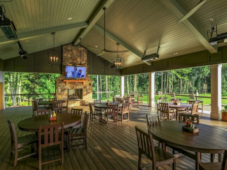 A spacious outdoor patio with wooden tables and chairs, a fireplace, and a TV, overlooking lush green trees and a grassy area.