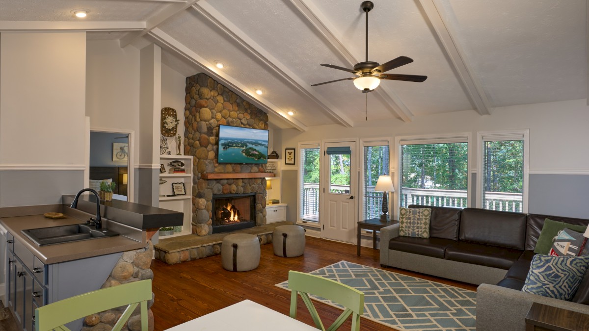 A cozy living room with a stone fireplace, ceiling fan, large windows, and modern furniture.