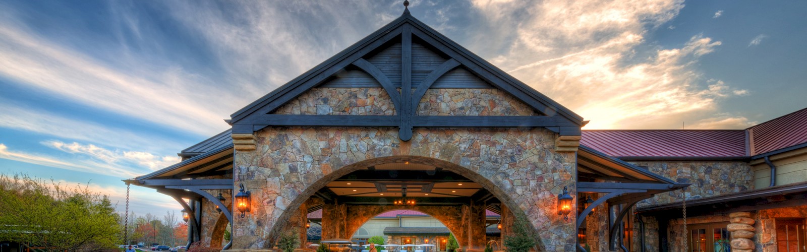 A stone archway entrance to a rustic building, with a well-landscaped area and dramatic clouds in the sky, during sunset.
