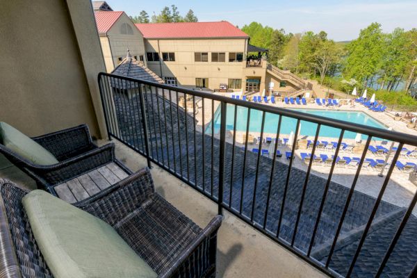A view from a balcony with two wicker chairs overlooking an outdoor swimming pool area, chairs with blue cushions, and surrounding trees.