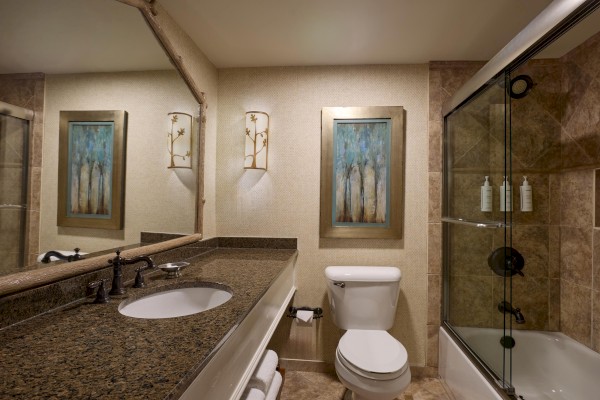 This image shows a bathroom with a sink, toilet, bathtub with sliding glass doors, wall art, and granite countertop.