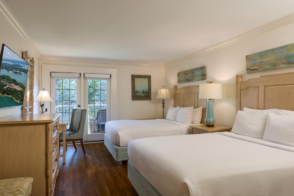 The image shows a well-lit hotel room with two beds, a dresser, lamps, wall art, and a door leading to a balcony with views.
